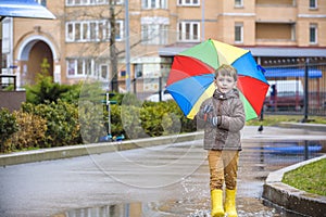 Little boy playing in rainy summer park. Child with colorful rainbow umbrella, waterproof coat and boots jumping in puddle and mud