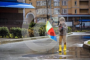 Little boy playing in rainy summer park. Child with colorful rainbow umbrella, waterproof coat and boots jumping in puddle and mud