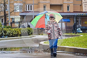 Little boy playing in rainy summer park. Child with colorful rainbow umbrella, waterproof coat and boots jumping in puddle and mud