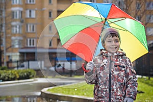 Little boy playing in rainy summer park. Child with colorful rainbow umbrella, waterproof coat and boots jumping in puddle and mud