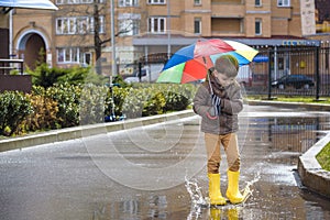 Little boy playing in rainy summer park. Child with colorful rainbow umbrella, waterproof coat and boots jumping in puddle and mud