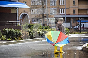 Little boy playing in rainy summer park. Child with colorful rainbow umbrella, waterproof coat and boots jumping in puddle and mud