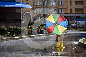 Little boy playing in rainy summer park. Child with colorful rainbow umbrella, waterproof coat and boots jumping in puddle and mud