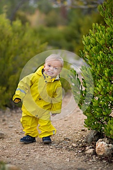 Little boy playing in rainy summer park. Child with colorful rainbow umbrella, waterproof coat and boots jumping in