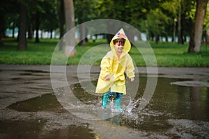 Little boy playing in rainy summer park. Child with umbrella, waterproof coat and boots jumping in puddle and mud in the