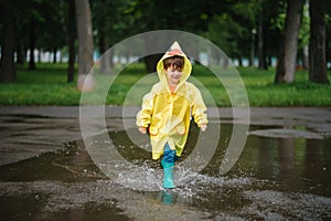 Little boy playing in rainy summer park. Child with umbrella, waterproof coat and boots jumping in puddle and mud in the