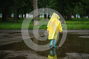 Little boy playing in rainy summer park. Child with umbrella, waterproof coat and boots jumping in puddle and mud in the