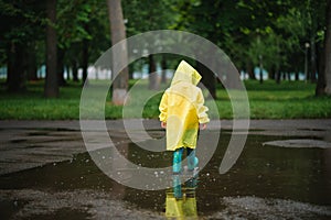 Little boy playing in rainy summer park. Child with umbrella, waterproof coat and boots jumping in puddle and mud in the