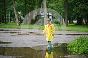 Little boy playing in rainy summer park. Child with umbrella, waterproof coat and boots jumping in puddle and mud in the