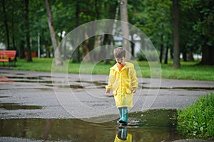 Little boy playing in rainy summer park. Child with umbrella, waterproof coat and boots jumping in puddle and mud in the