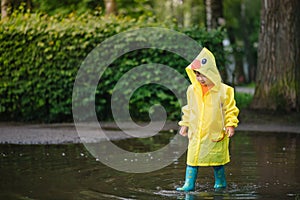 Little boy playing in rainy summer park. Child with umbrella, waterproof coat and boots jumping in puddle and mud in the