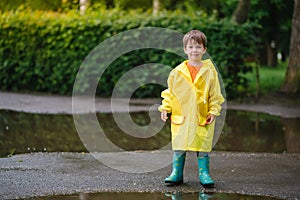 Little boy playing in rainy summer park. Child with umbrella, waterproof coat and boots jumping in puddle and mud in the