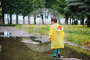 Little boy playing in rainy summer park. Child with umbrella, waterproof coat and boots jumping in puddle and mud in the