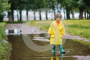 Little boy playing in rainy summer park. Child with umbrella, waterproof coat and boots jumping in puddle and mud in the