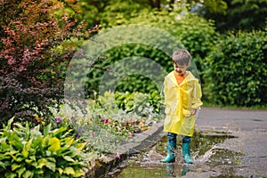 Little boy playing in rainy summer park. Child with umbrella, waterproof coat and boots jumping in puddle and mud in the