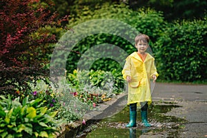 Little boy playing in rainy summer park. Child with umbrella, waterproof coat and boots jumping in puddle and mud in the