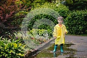 Little boy playing in rainy summer park. Child with umbrella, waterproof coat and boots jumping in puddle and mud in the