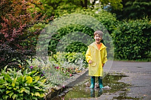 Little boy playing in rainy summer park. Child with umbrella, waterproof coat and boots jumping in puddle and mud in the