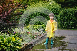 Little boy playing in rainy summer park. Child with umbrella, waterproof coat and boots jumping in puddle and mud in the