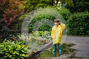 Little boy playing in rainy summer park. Child with umbrella, waterproof coat and boots jumping in puddle and mud in the