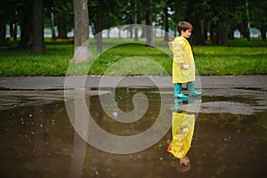 Little boy playing in rainy summer park. Child with umbrella, waterproof coat and boots jumping in puddle and mud in the