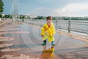 Little boy playing in rainy summer park. Child with umbrella, waterproof coat and boots jumping in puddle and mud in the