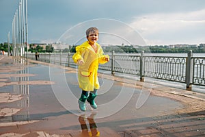 Little boy playing in rainy summer park. Child with umbrella, waterproof coat and boots jumping in puddle and mud in the