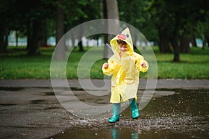 Little boy playing in rainy summer park. Child with umbrella, waterproof coat and boots jumping in puddle and mud in the rain. Kid