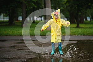 Little boy playing in rainy summer park. Child with umbrella, waterproof coat and boots jumping in puddle and mud in the rain. Kid