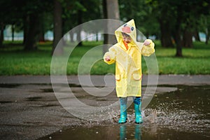 Little boy playing in rainy summer park. Child with umbrella, waterproof coat and boots jumping in puddle and mud in the rain. Kid