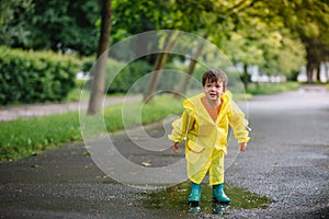 Little boy playing in rainy summer park. Child with umbrella, waterproof coat and boots jumping in puddle and mud in the rain. Kid