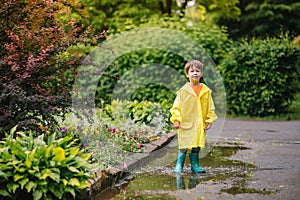 Little boy playing in rainy summer park. Child with umbrella, waterproof coat and boots jumping in puddle and mud in the rain. Kid