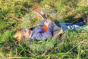 A little boy plays the guitar while lying on the green grass in the park