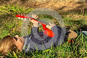 A little boy plays the guitar while lying on the green grass in the park