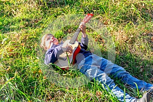 A little boy plays the guitar while lying on the green grass in the park