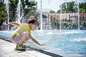 Little boy plays in the square near pool with water jets in the fountain at sunny summer day. Active summer leisure for kids in