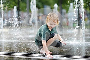 Little boy plays in the square between the water jets in the city fountain at sunny summer day. Active summer leisure for kids in