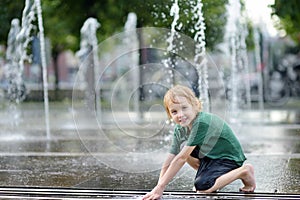 Little boy plays in the square between the water jets in the city fountain at sunny summer day. Active summer leisure for kids in