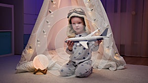 Little boy plays with toy white airplane, dreams of flying and being captain of an aircraft sitting in dark room near