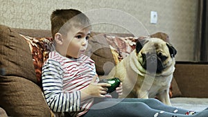 Little boy plays in video game with joystick, pug dog with child on the couch at home