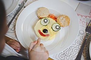 Little boy pointing his small finger at plate with funny tasty dish with cartoon character face from food