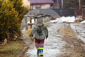 Little boy in protective rubber boots and rain clothes jumping in mud puddle. Happy child having fun while playing in