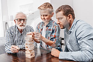 Little boy pulling a piece out of blocks wood tower while his father and grandfather