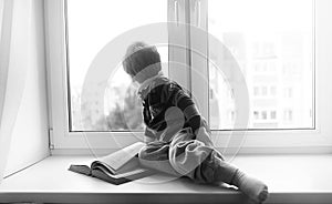 The little boy is reading a book. The child sits at the window a