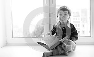 The little boy is reading a book. The child sits at the window a