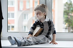 The little boy is reading a book. The child sits at the window a