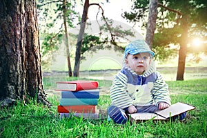 Little boy reading a book while sitting on the green grass in park. Stack of multicolored textbooks and cute baby.