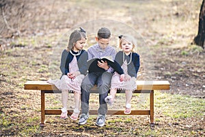 Little boy reading a book for two girls sitting on a bench in a park