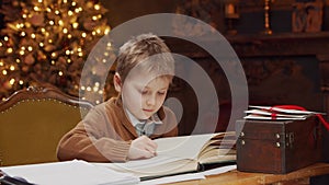 Little boy reads a magic book while sitting at the table. Home interior with Christmas tree and fireplace. Christmas