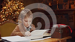 Little boy reads a magic book while sitting at the table. Home interior with Christmas tree and fireplace. Christmas